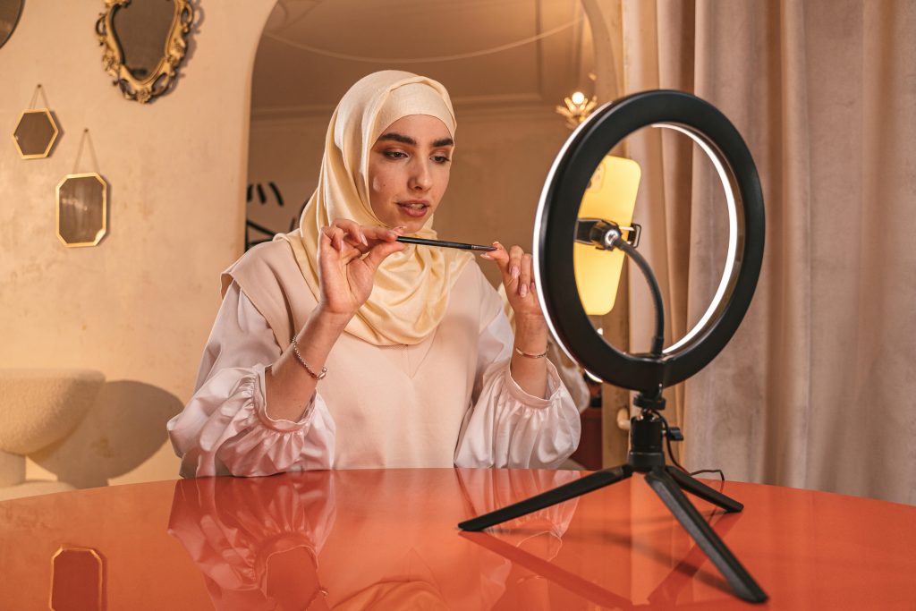 Muslim woman using ring light for creating online makeup content indoors.